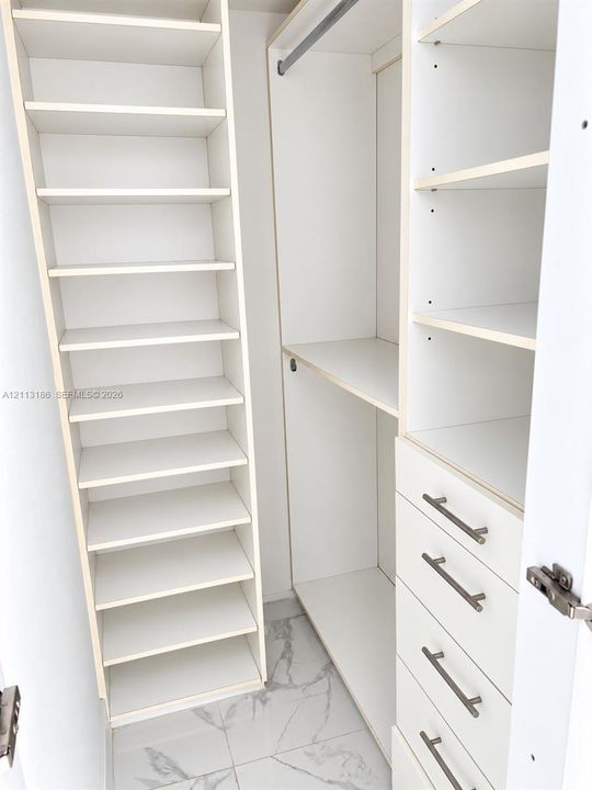 Custom closet organization system featuring floor-to-ceiling shelving, hanging rods, and four built-in drawers with metallic hardware finished with stone-patterned tile flooring