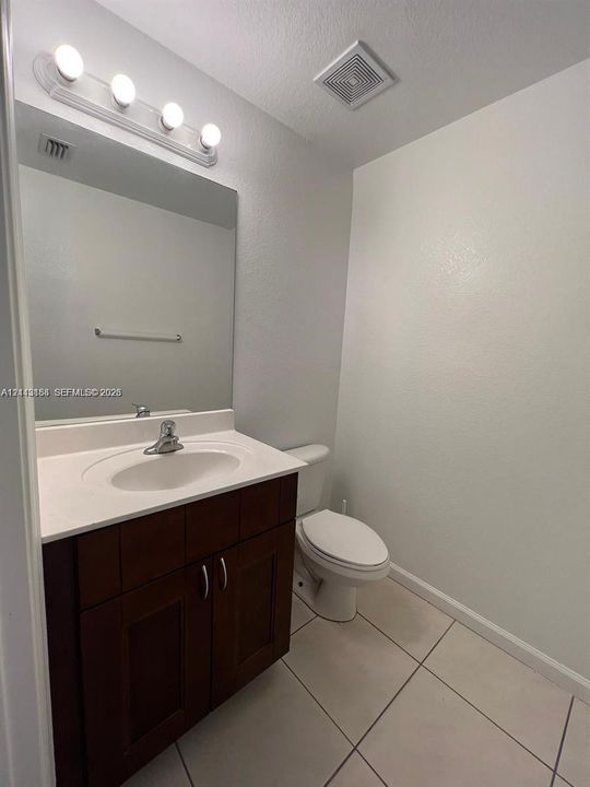 Dark wood-finish vanity with a white countertop, integrated sink, polished chrome faucet, and large wall-mounted vanity mirror with multi-bulb light fixture, complemented by tile flooring