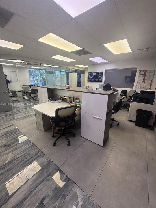 Interior workspace featuring large-format ceramic floor tiling, glass partition walls, built-in cabinetry with integrated desk surfaces, and a drop ceiling with recessed lighting
