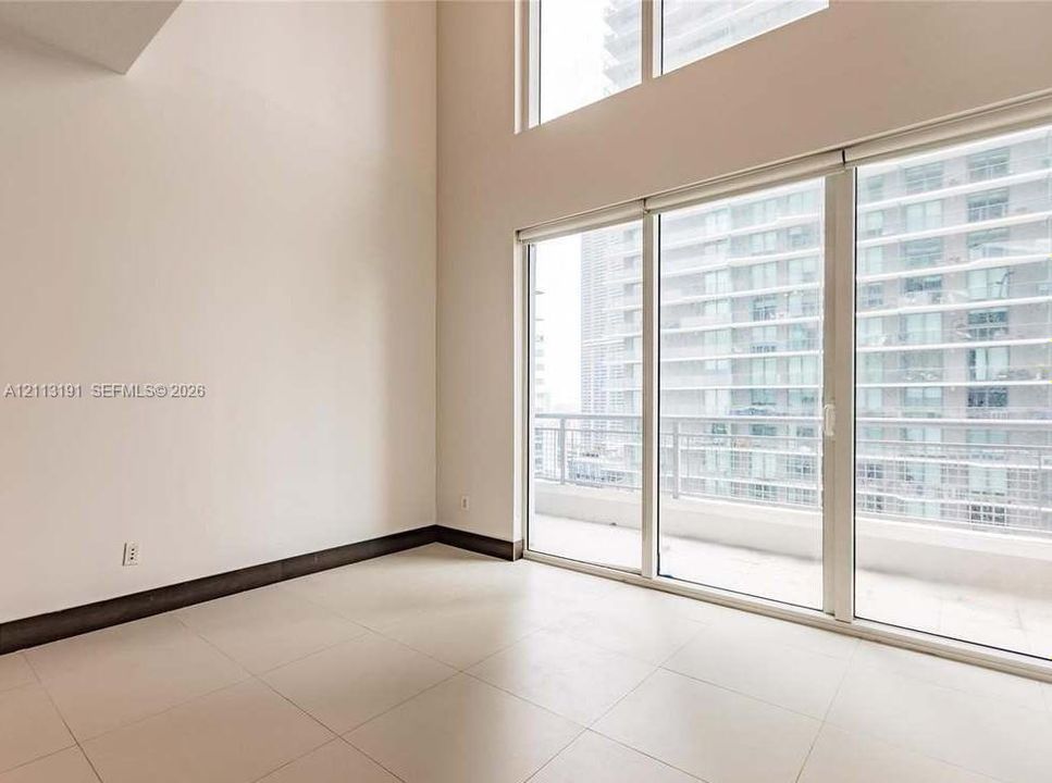 Double-height ceiling, floor-to-ceiling windows, light-toned tiled flooring, and sliding glass doors opening to a private balcony