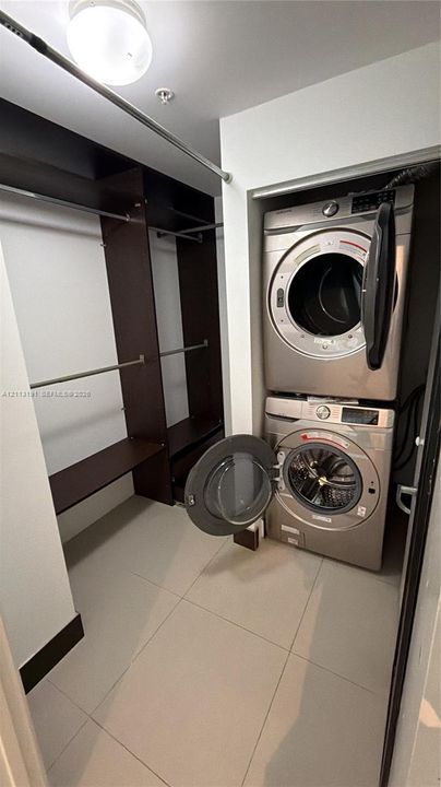 Utility area featuring stacked laundry appliances, large-format tile flooring, and integrated dark wood-finish shelving with built-in clothing rods