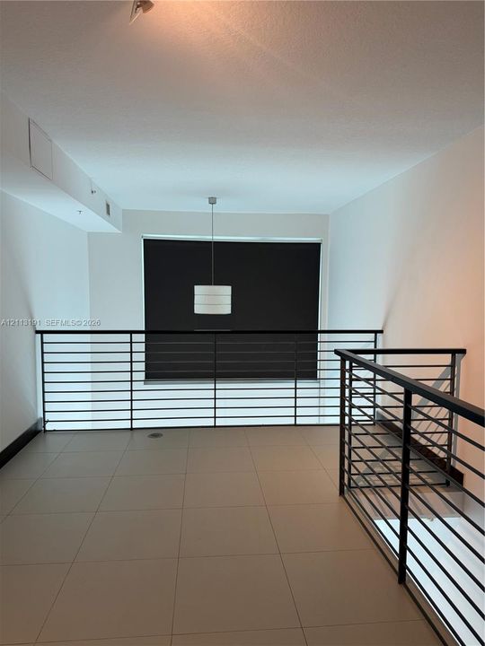 Open-concept mezzanine area featuring large-format tile flooring, industrial-style black horizontal metal railings, and a suspended drum pendant light fixture
