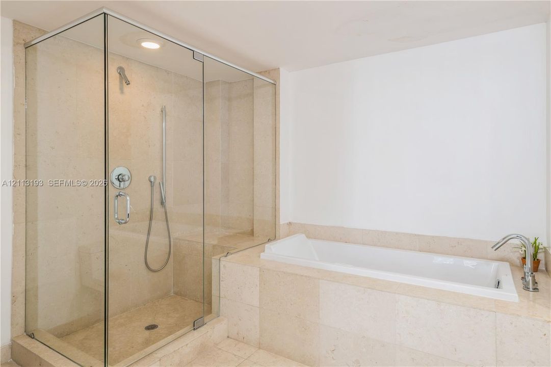 Walk-in glass-enclosed shower with stone-finish tiling and built-in bench, paired with a drop-in soaking tub featuring stone-finish surround and metal faucet hardware