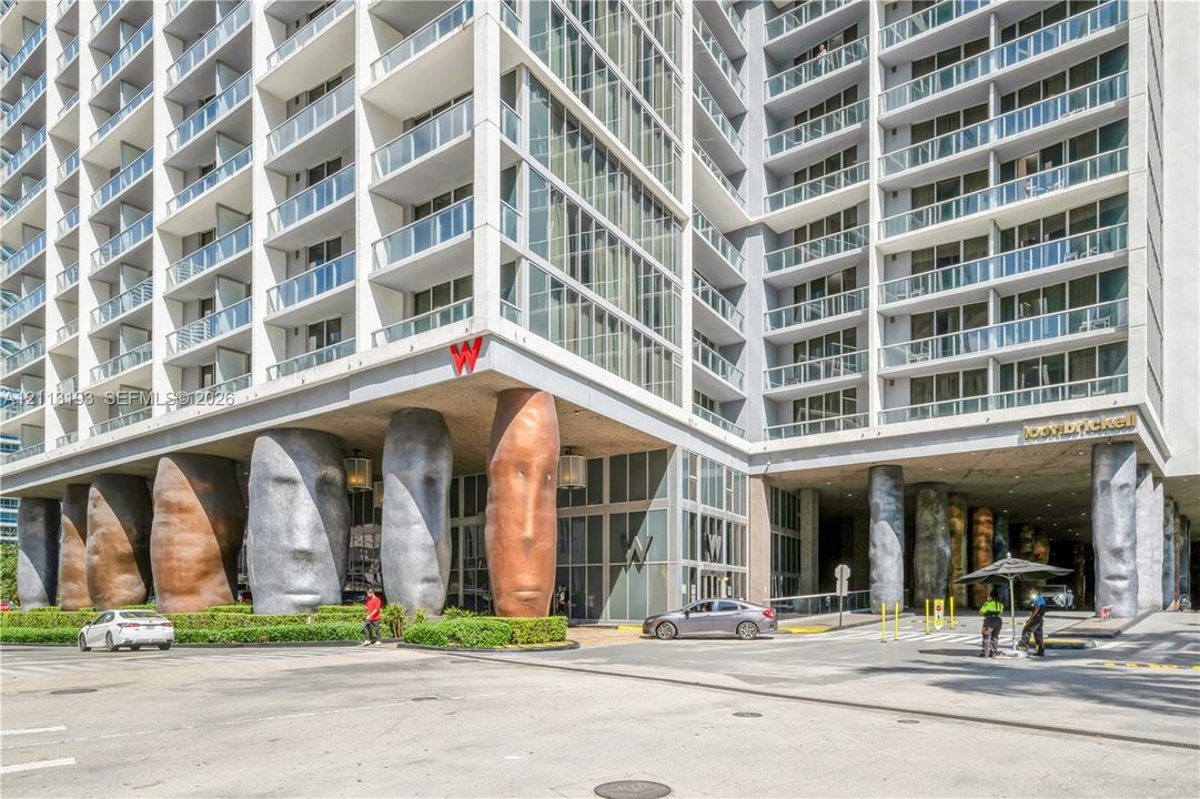 High-rise exterior featuring a multi-story glass facade, private glass-railed balconies, and distinctive sculpted structural columns at the building entrance