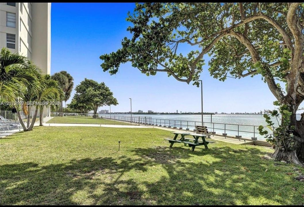 Waterfront grounds featuring mature canopy trees, a manicured lawn, metal safety railing, and paved perimeter pathways