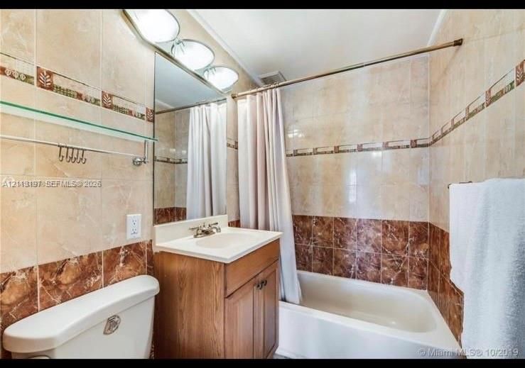 Full bathroom featuring floor-to-ceiling tile, a wood-finish vanity with an integrated sink, a deep soaking tub, and a decorative tile inlay border