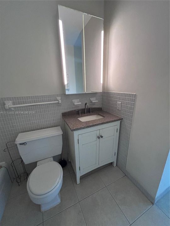 Bathroom featuring a vanity with stone-finish countertop, under-mount sink, white cabinetry, integrated medicine cabinet with built-in vertical lighting, and tiled wall surround