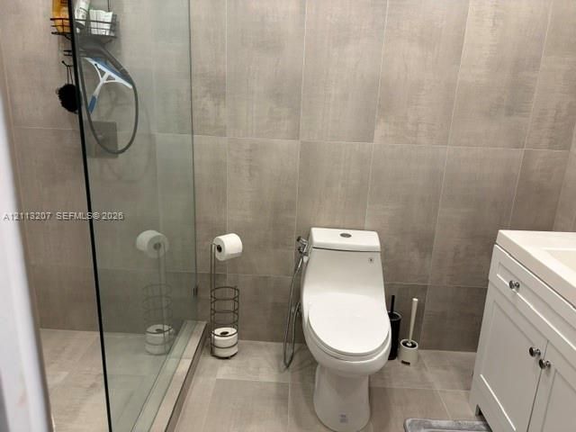 Bathroom featuring floor-to-ceiling large-format neutral tile, a frameless glass-enclosed shower, a white shaker-style vanity with integrated countertop, and a dual-flush toilet