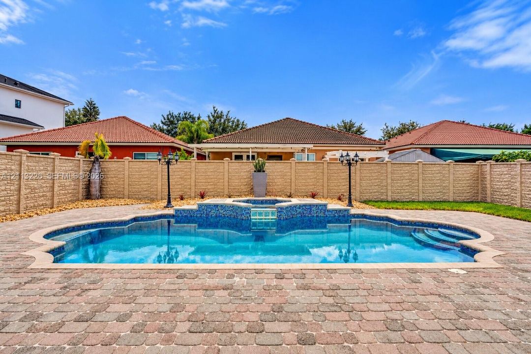 Custom-shaped in-ground swimming pool featuring a tiled spa with waterfall element, surrounded by paver decking and enclosed by a textured masonry privacy wall