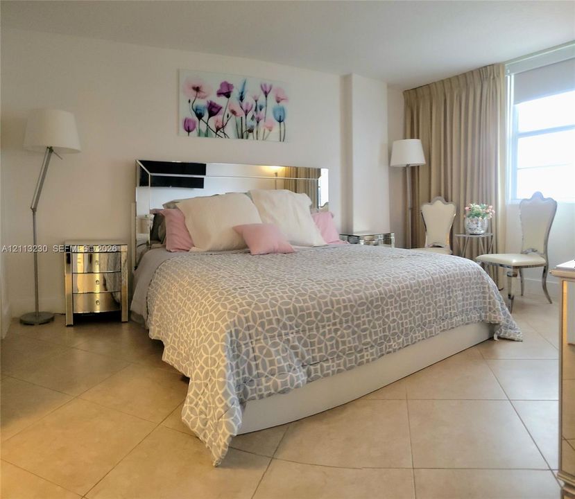Large-format neutral tile flooring, mirrored wall-mounted headboard, and floor-to-ceiling window treatment