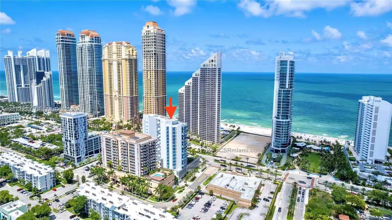 Aerial perspective of a high-rise residential building featuring a glass-facade exterior, multiple balconies, and a ground-level swimming pool, situated in a coastal location