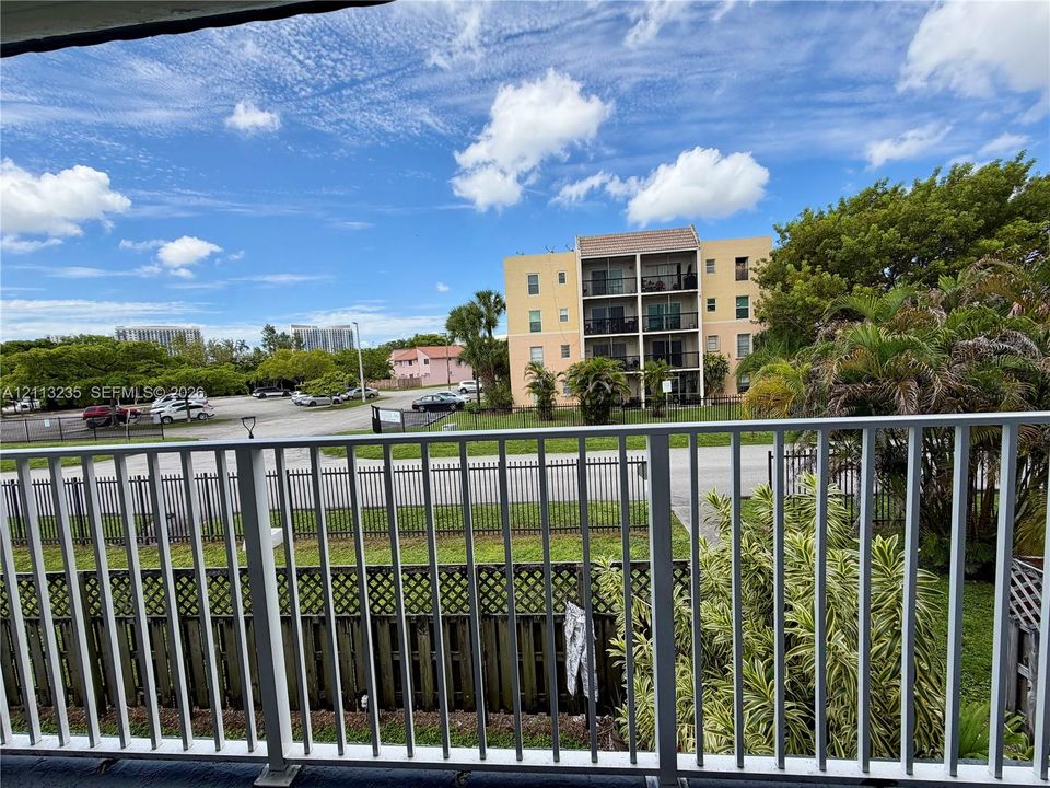 Elevated balcony featuring a white metal railing, overlooks a grassy lawn, mature landscaping, and surrounding community infrastructure