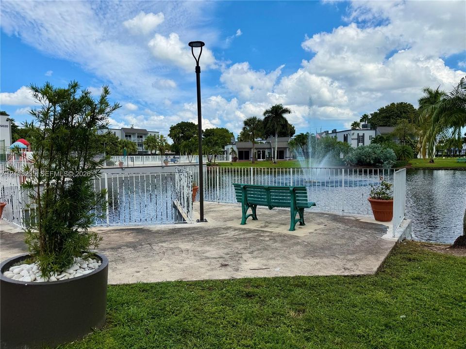 Paved waterfront terrace featuring a decorative fountain, metal perimeter fencing, and mature landscaping