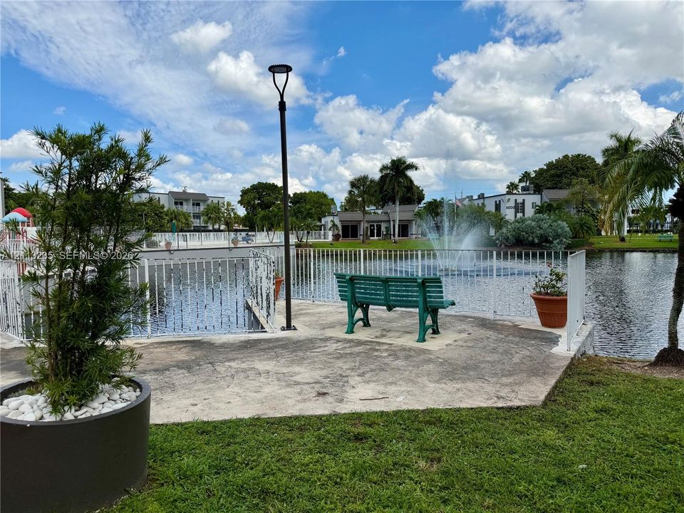 Concrete patio area featuring a water fountain, white metal fencing, a lamp post, and professional landscaping