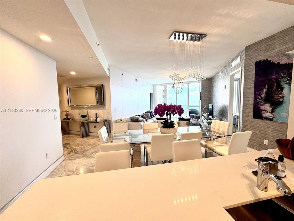 Open floor plan featuring polished stone flooring, custom textured wall cladding, floor-to-ceiling windows, and a modern tiered crystal chandelier