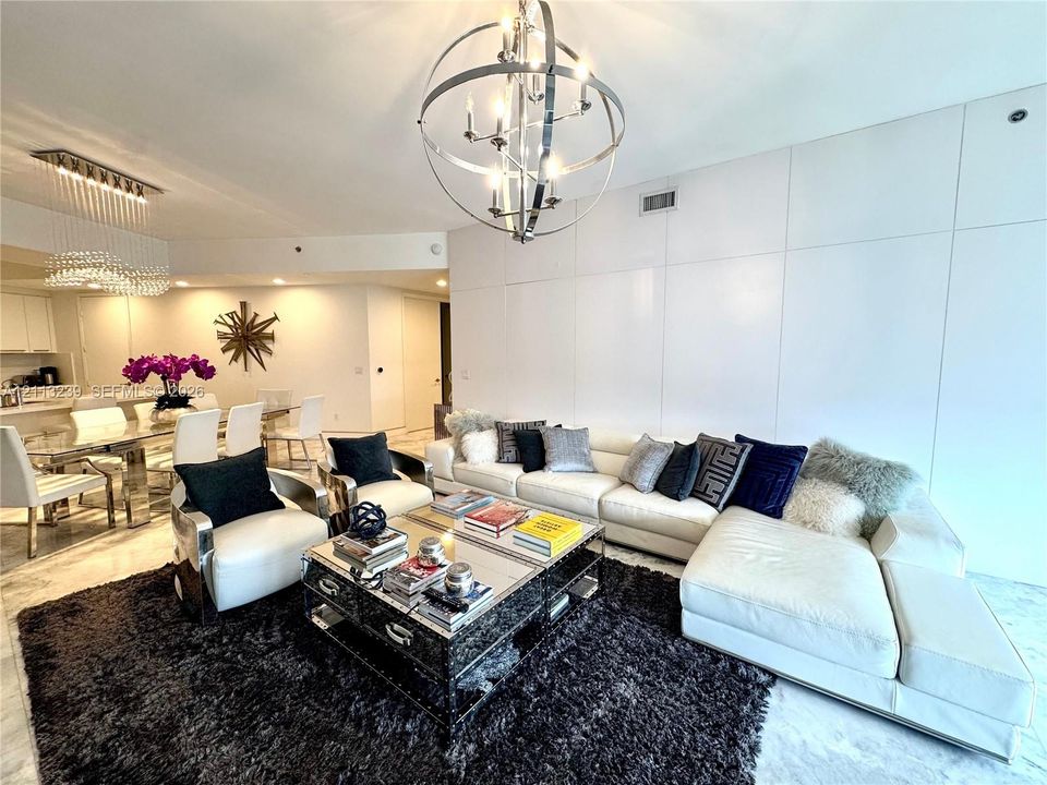 Open-concept living space featuring high-gloss, full-height wall paneling, polished stone flooring, a multi-arm spherical chandelier, and a linear crystal ceiling fixture