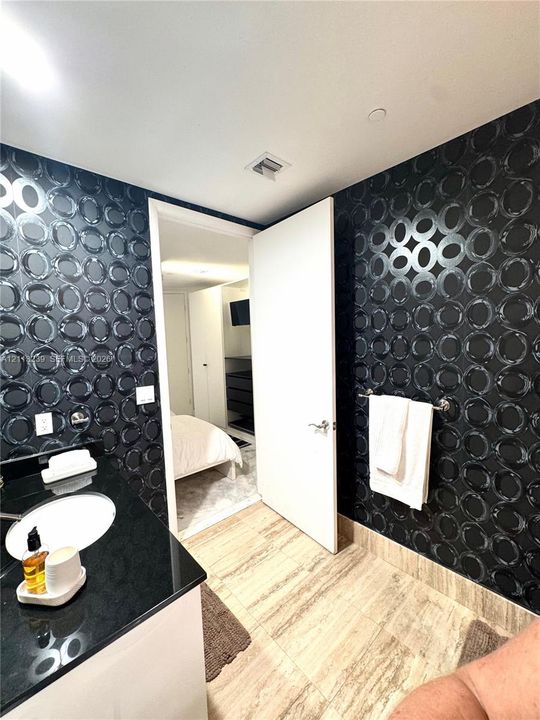 Bathroom featuring high-contrast geometric patterned wall covering, stone-finish floor tile, and a black countertop vanity with a white drop-in basin