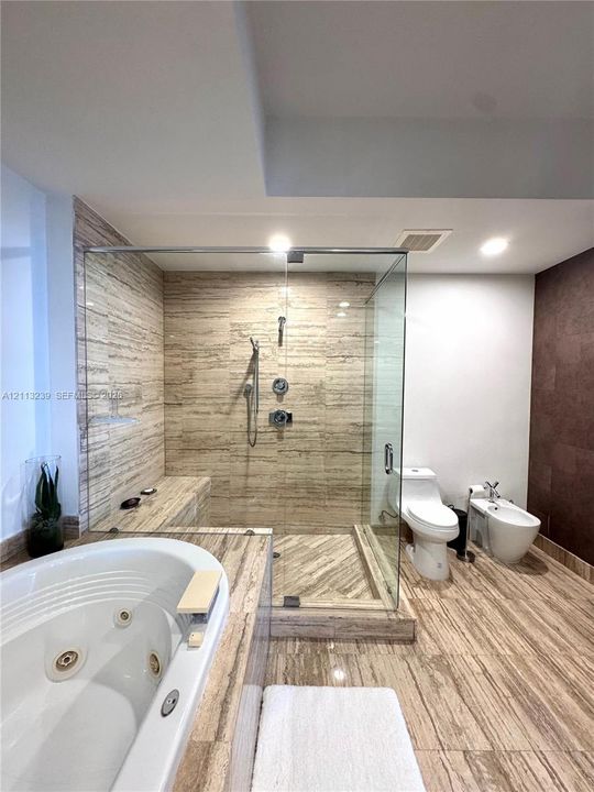 Full-size jetted soaking tub, walk-in shower with stone-tile surround, built-in shower bench, and stone-tile flooring