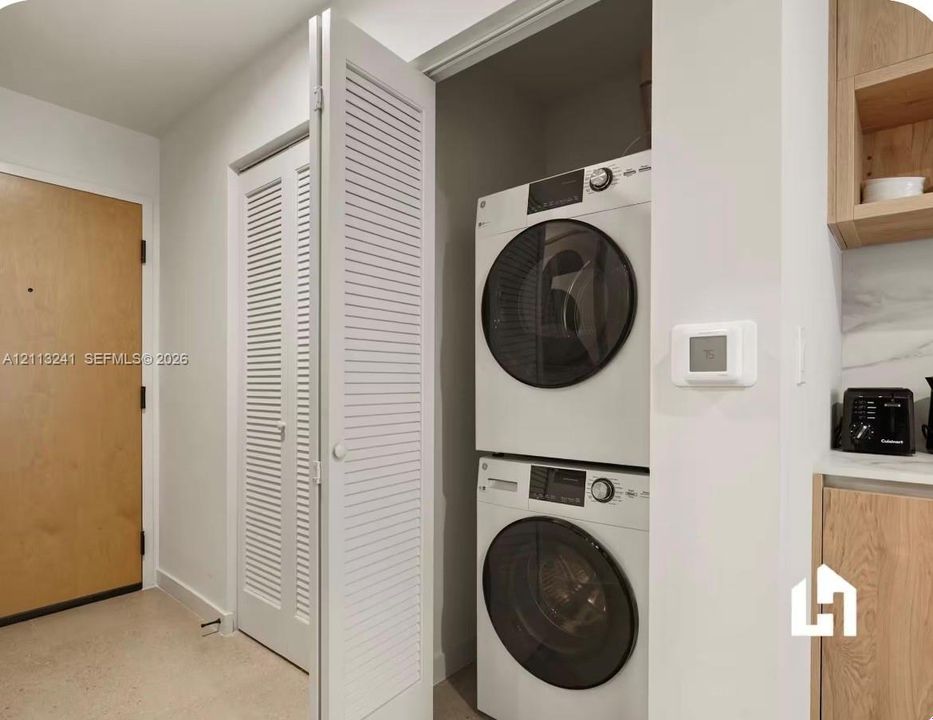 Stacked laundry center with white front-loading appliances housed in a louvered-door closet, featuring a smart thermostat and light-wood cabinetry with stone-surface counters