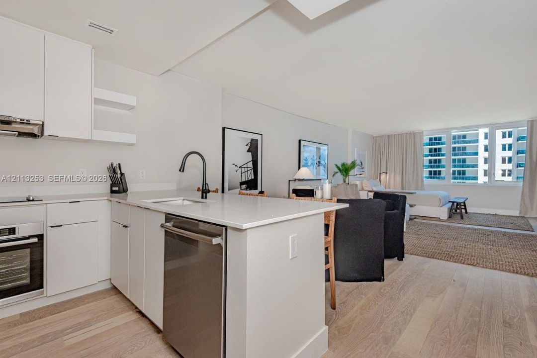 Kitchen featuring white flat-panel cabinetry, light-toned stone countertops, a stainless steel dishwasher, a matte black high-arch faucet, and light wood-finish flooring