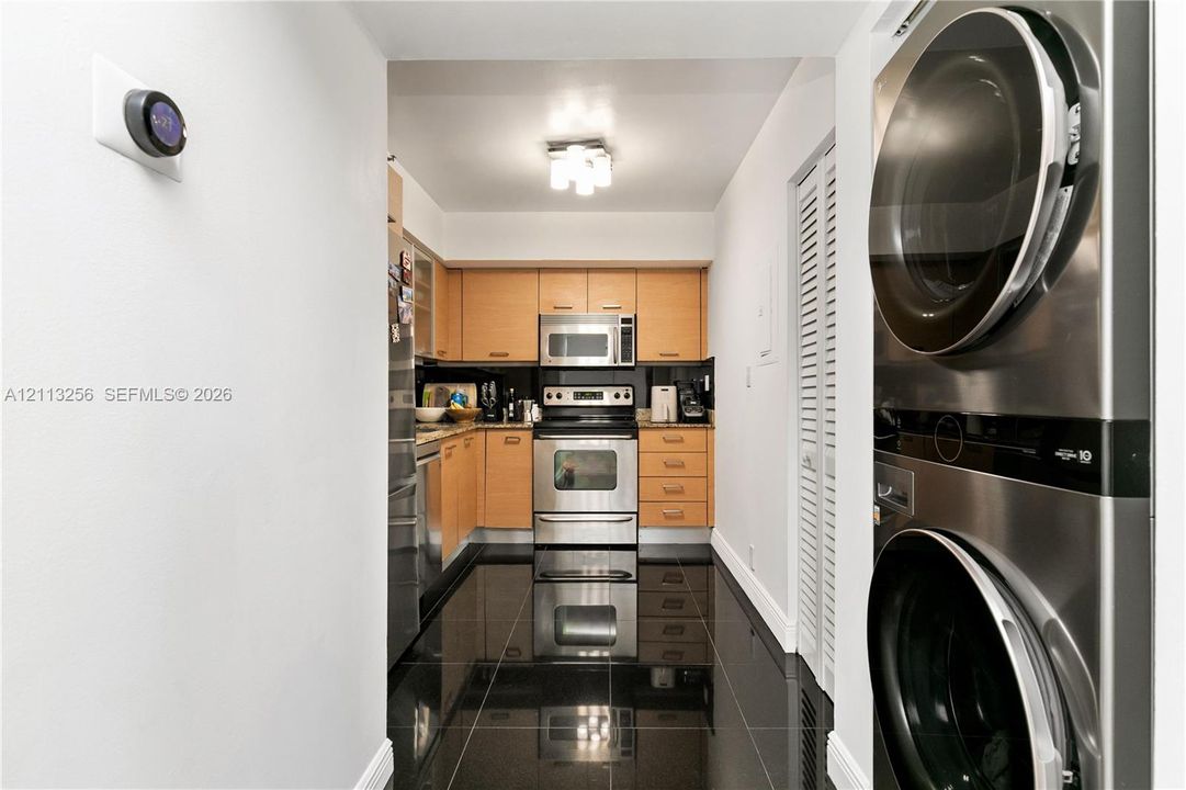 Kitchen featuring wood-finish cabinetry, stainless steel appliances, dark tile flooring, and a stacked laundry unit with louvered closet doors
