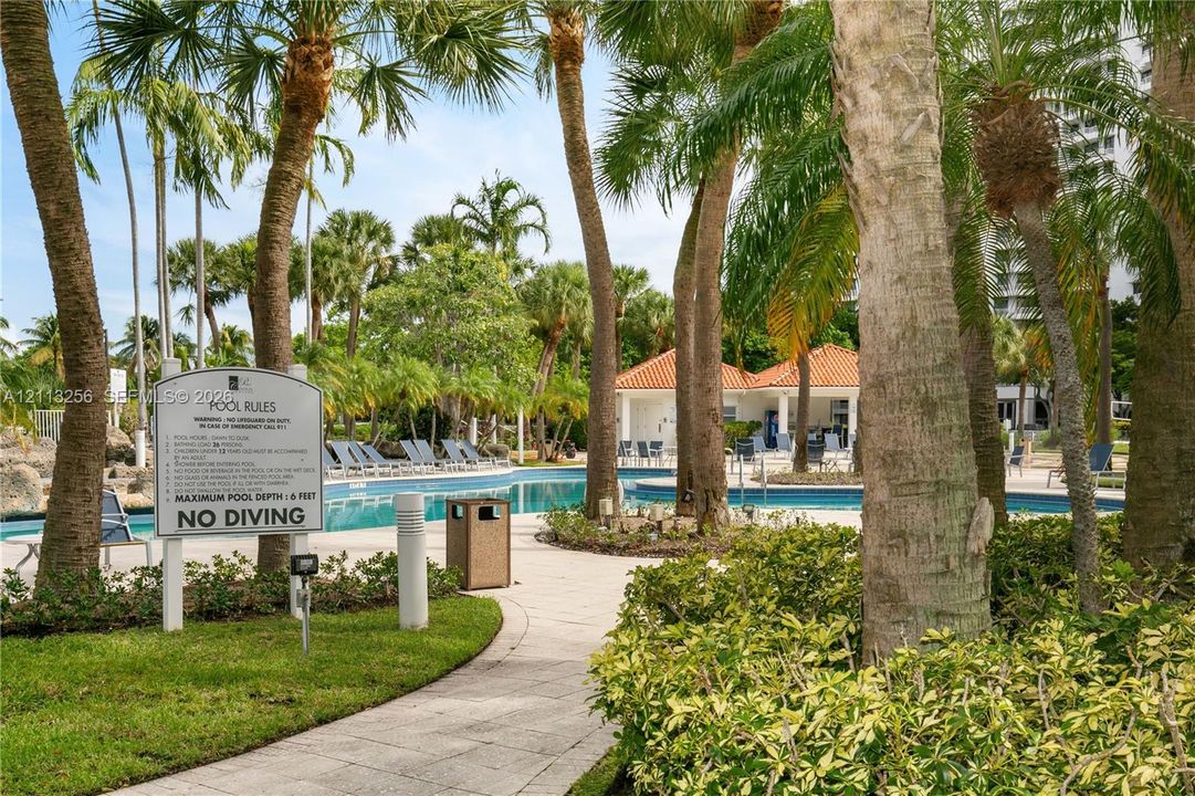 Swimming pool area featuring a stone-paved deck, mature palm trees, landscaped shrubbery, and a covered structure with a terracotta tile roof