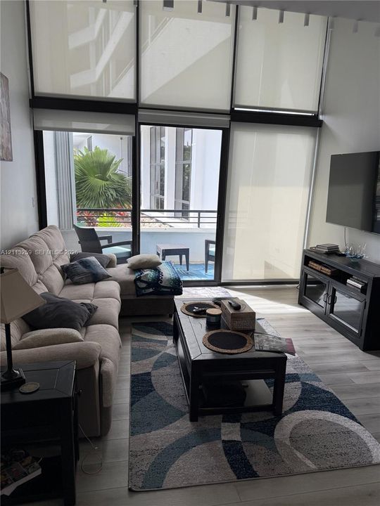 Floor-to-ceiling windows with roller shades, wood-finish flooring, and sliding glass door opening to a private balcony