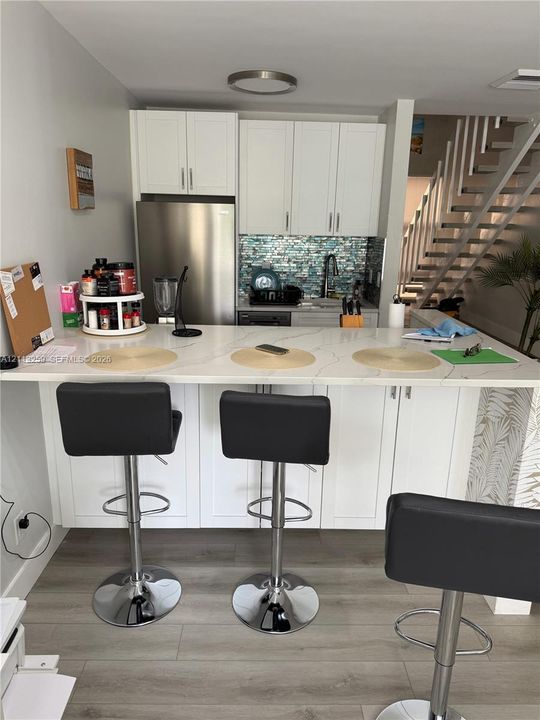 Kitchen featuring white shaker-style cabinetry, a stone-finish countertop peninsula, mosaic tile backsplash, stainless steel refrigerator, and wood-finish flooring