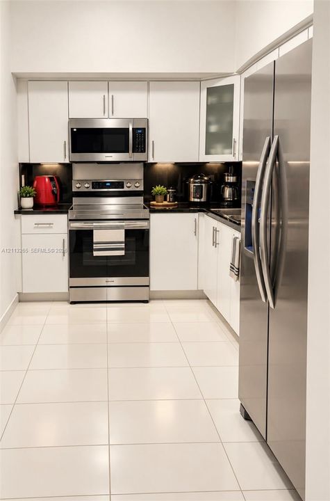 Kitchen featuring white shaker-style cabinetry, dark stone countertops, stainless steel appliances, a glass-front upper cabinet, and light-toned tile flooring