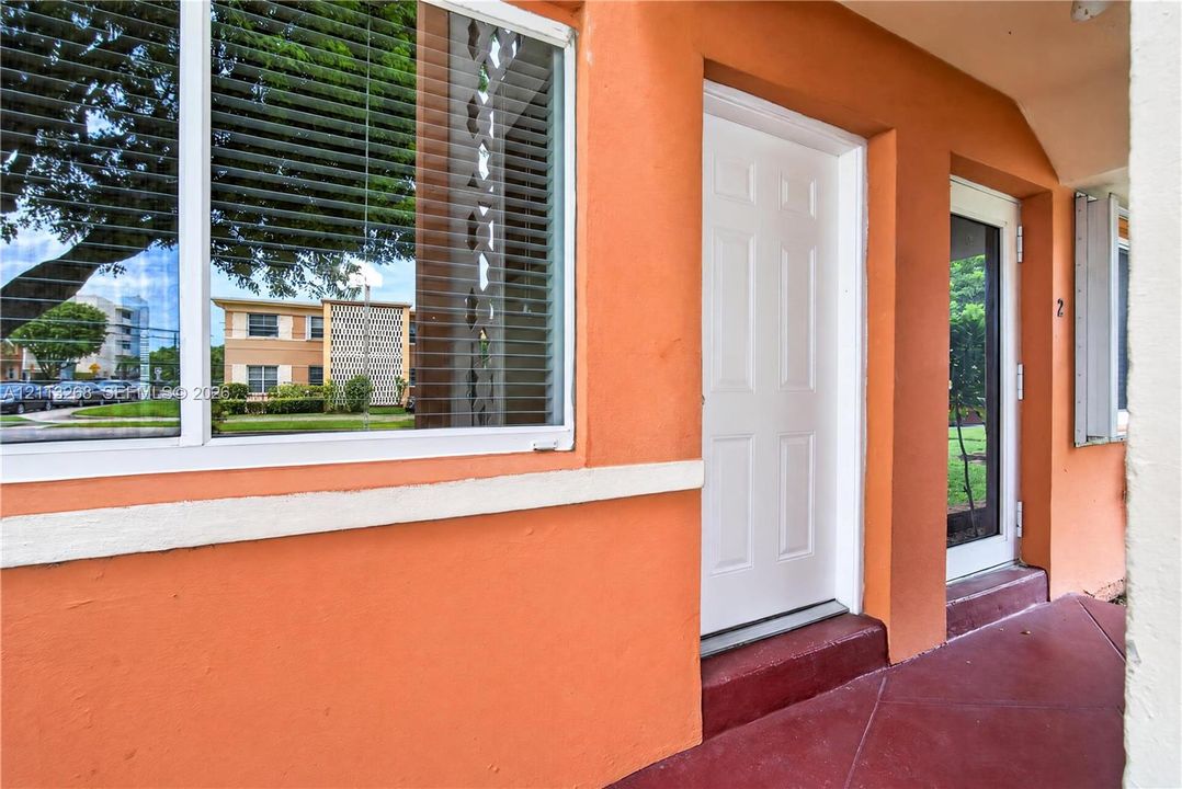Stucco exterior featuring a large window with white framing, solid white entry door, glass-paneled storm door, and concrete flooring