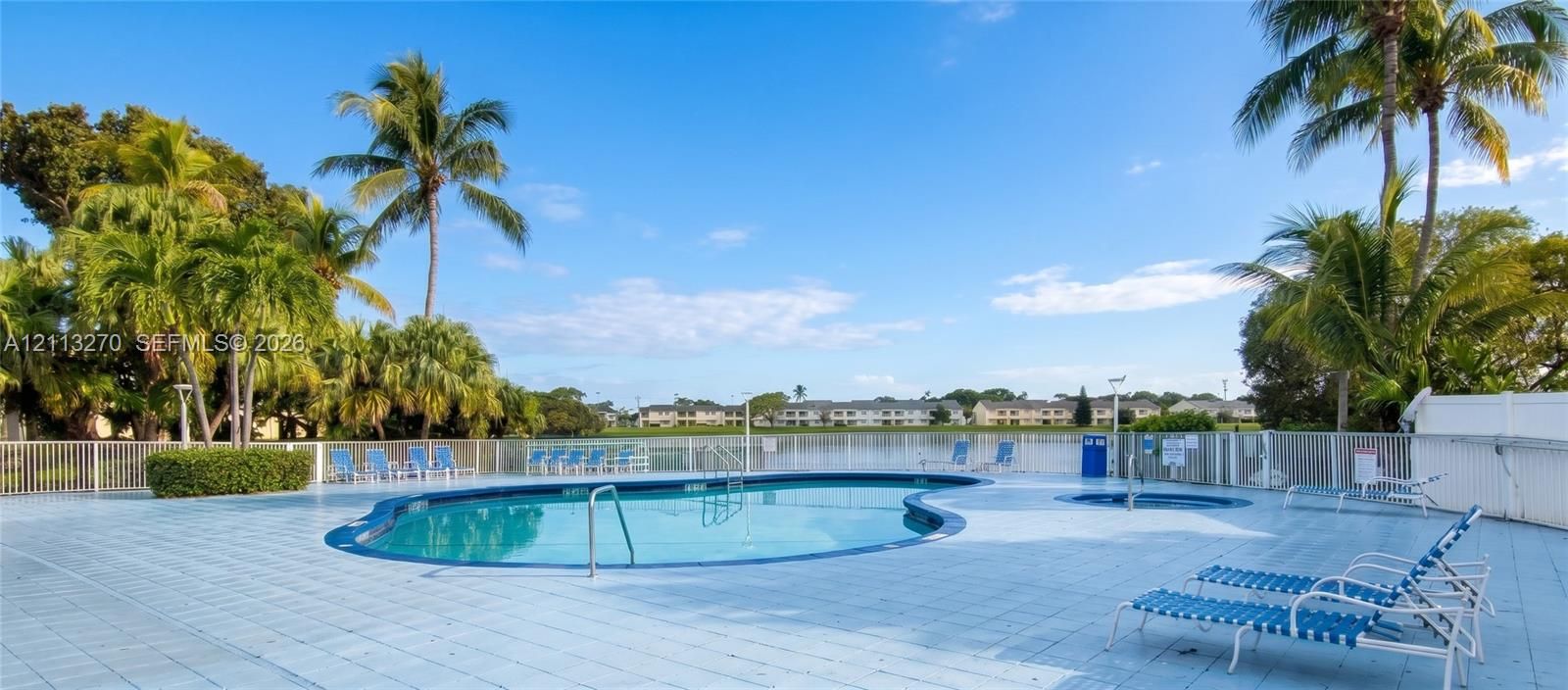 In-ground swimming pool and circular spa featuring a paved deck, perimeter fencing, and mature palm trees overlooking a lake
