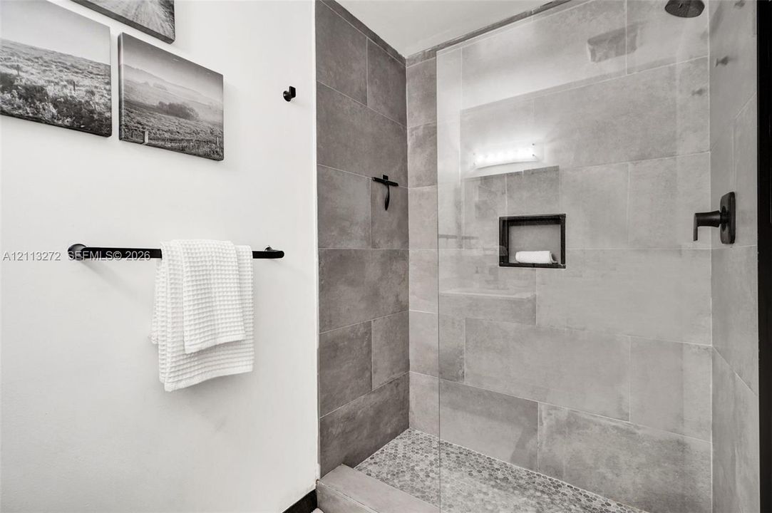 Walk-in shower featuring large-format grey tile surround, pebble-texture floor, frameless glass enclosure, built-in recessed wall niche, and matte black plumbing fixtures