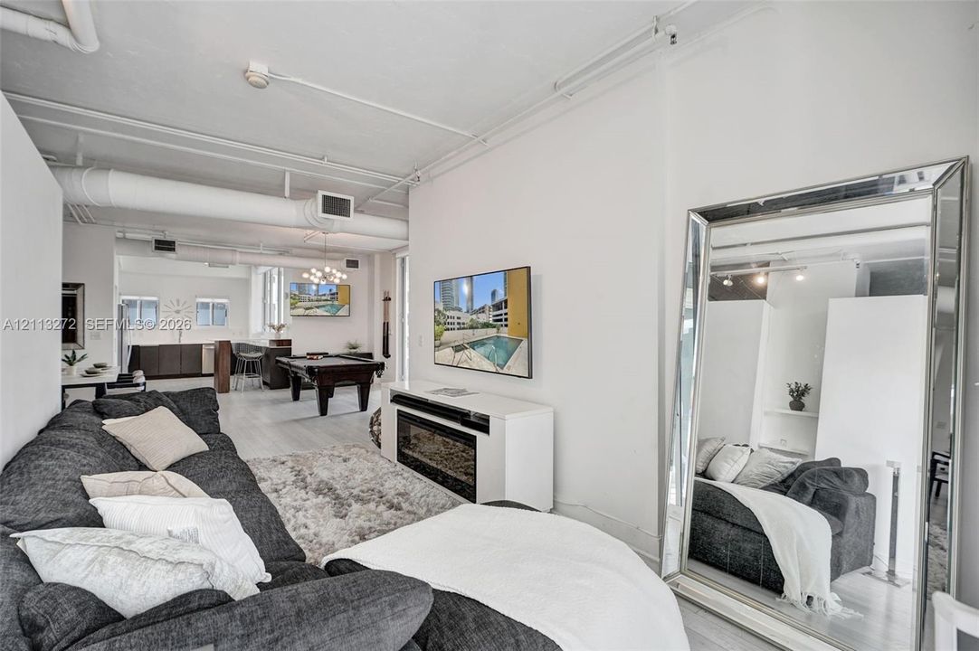 Open-concept interior featuring industrial-style exposed ductwork, white painted brick walls, wood-finish flooring, and a built-in wall-mounted electric fireplace