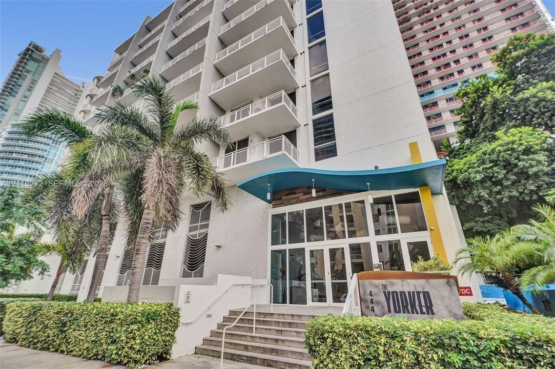 Multi-story high-rise exterior featuring concrete balconies, floor-to-ceiling glass-paneled ground-level entryway, decorative blue architectural awning, and stone-tiled stairs