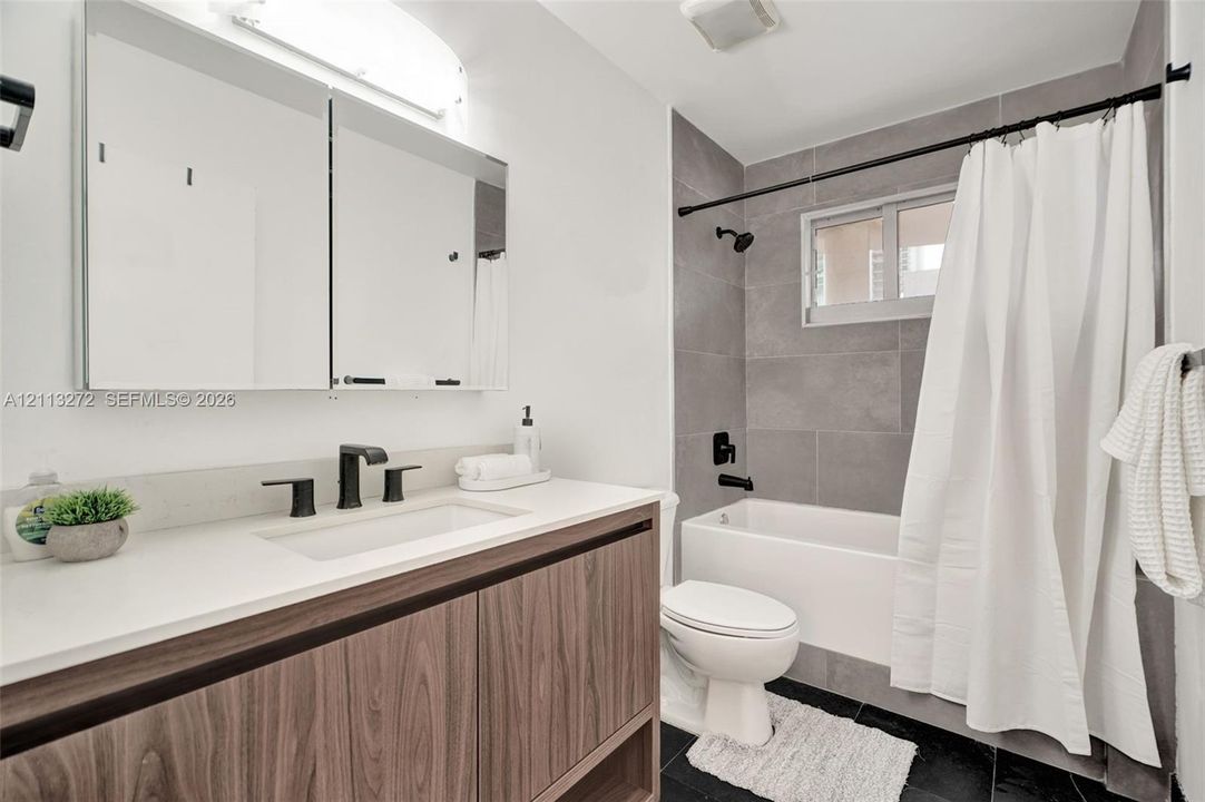 Bathroom featuring a wood-finish vanity with a white countertop, large mirrored medicine cabinet, matte black plumbing fixtures, and a tub-shower combination with grey tile surround