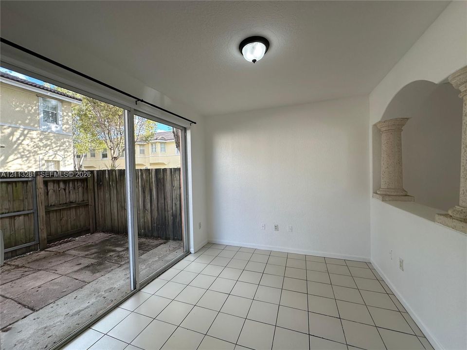 Interior space featuring light-colored tile flooring, decorative architectural columns, a ceiling-mounted light fixture, and a large sliding glass door opening to a concrete patio enclosed by wood fencing