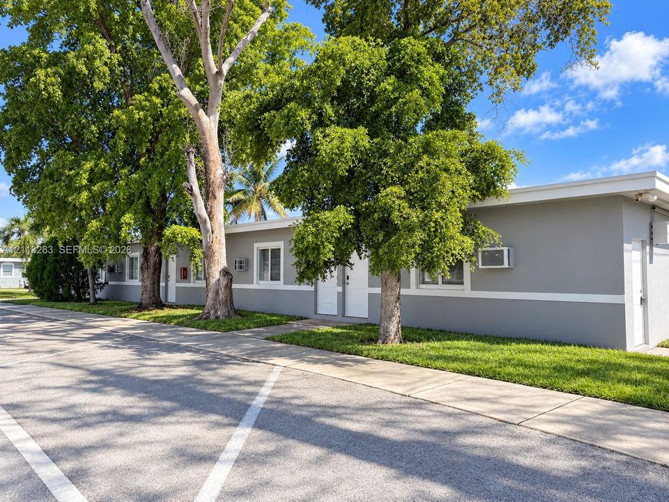 Single-story exterior featuring light gray painted masonry, white trim detailing, flat roofline, paved parking area, and mature trees
