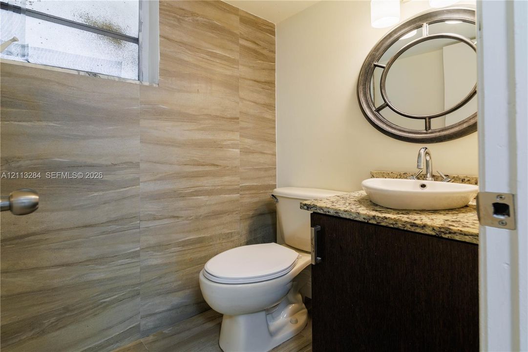 Vanity featuring a stone-look countertop, integrated oval vessel sink, and dark-finish cabinetry, complemented by wall-to-wall wood-tone tile and a circular decorative wall mirror
