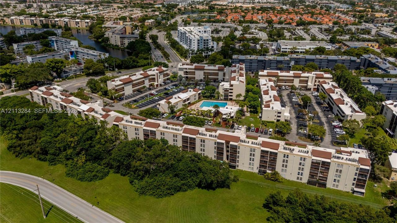 Aerial perspective of a multi-story residential complex featuring beige stucco facades, multi-pitched brown roofing, integrated parking areas, and a central outdoor swimming pool