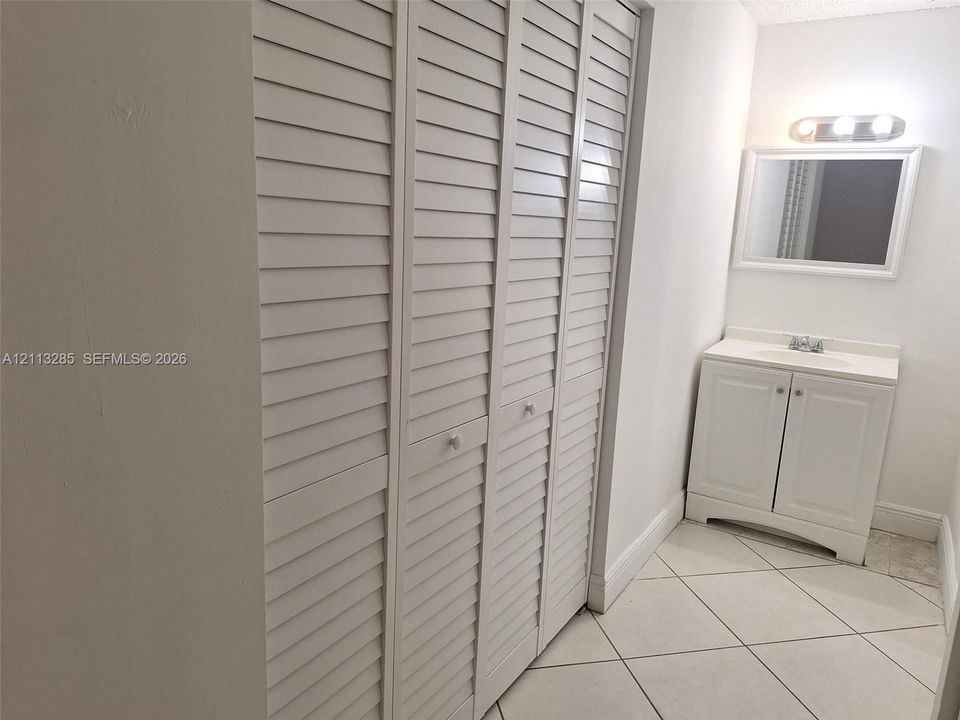 White louvered closet doors, vanity with integrated basin, polished light-colored tiled flooring, and wall-mounted vanity mirror with integrated lighting fixture