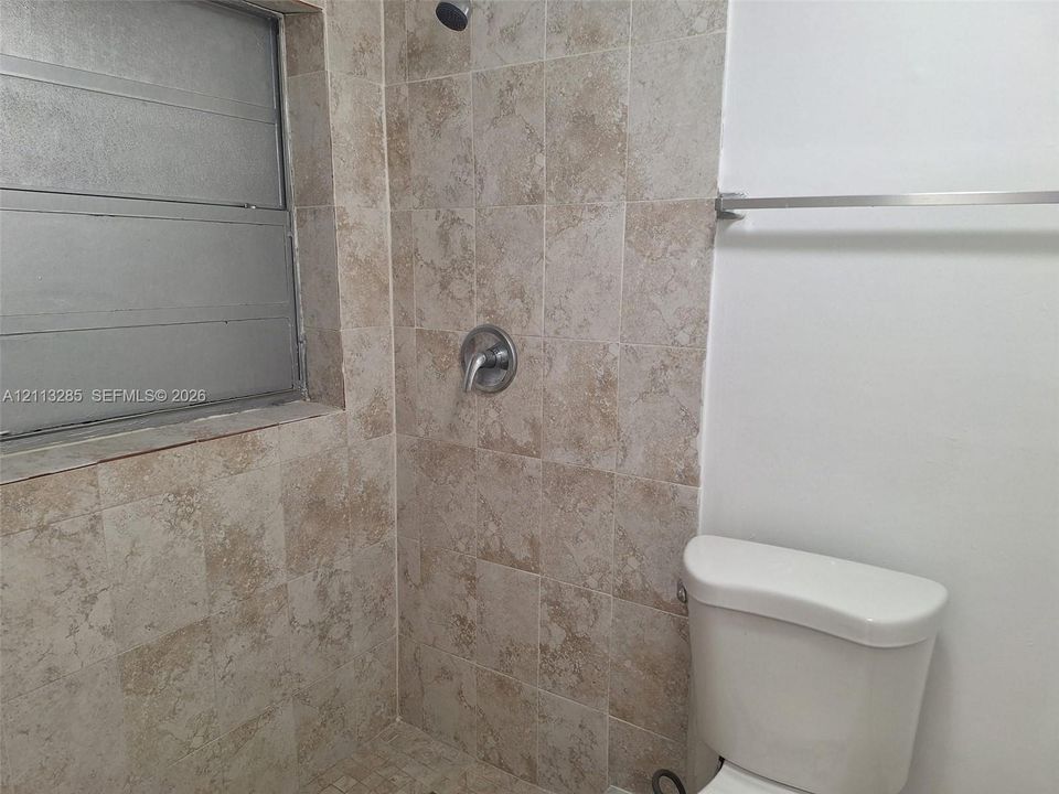 Shower enclosure featuring textured tile walls, a stainless steel single-handle shower faucet, a louvered window, and a wall-mounted metal towel bar