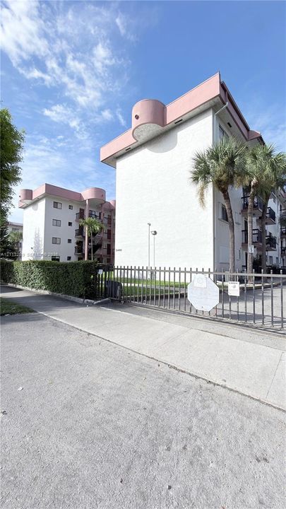 Multi-story residential buildings featuring white stucco facades, decorative roofline architectural accents, tiered balcony railings, and a secure metal perimeter gate