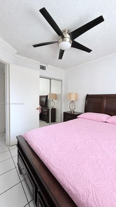 Room featuring white ceramic tile flooring, crown molding, a brushed nickel and black ceiling fan with integrated lighting, and a closet with mirrored sliding doors