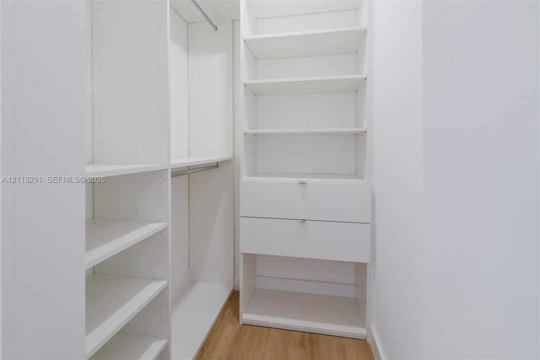 Built-in white storage system featuring adjustable shelving, integrated hanging rods, and dual drawers with silver-tone hardware over wood-finish flooring