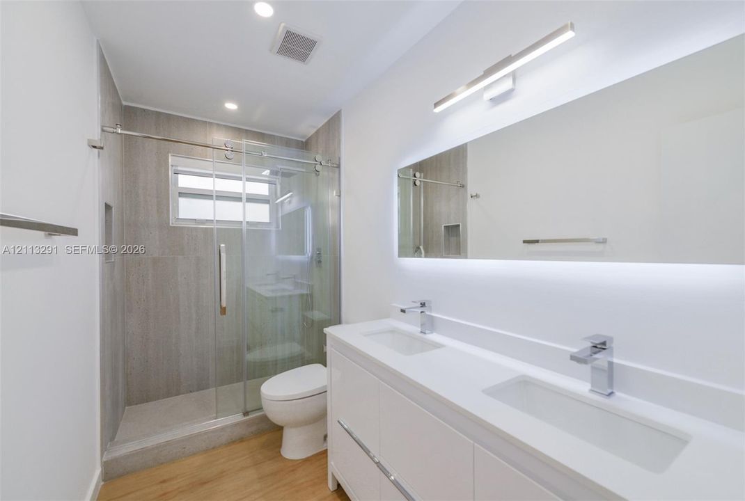 Double vanity with a white countertop and rectangular undermount sinks, illuminated mirror with integrated backlighting, wood-finish flooring, and a walk-in shower featuring sliding glass doors and neutral large-format tile surround