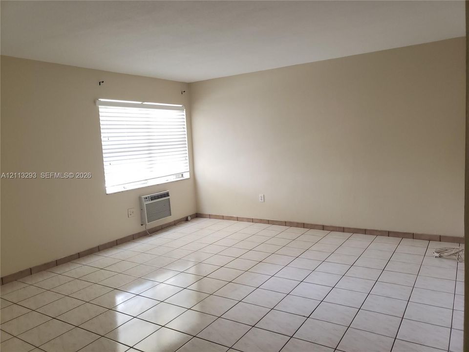 Interior room featuring light-colored ceramic tile flooring, neutral wall paint, a window with horizontal blinds, and a built-in wall-mounted air conditioning unit