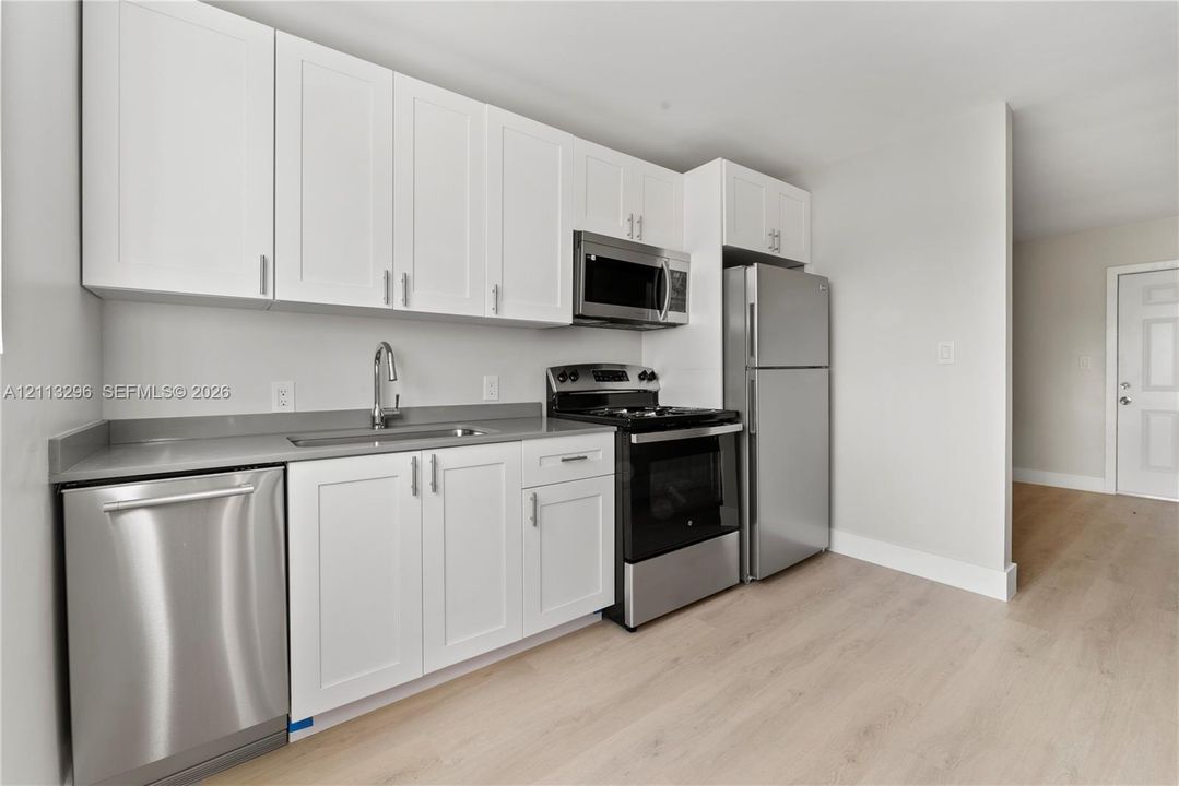 Kitchen featuring white shaker-style cabinetry, grey solid-surface countertops, wood-finish flooring, and a stainless steel appliance package including a dishwasher, microwave, range, and refrigerator