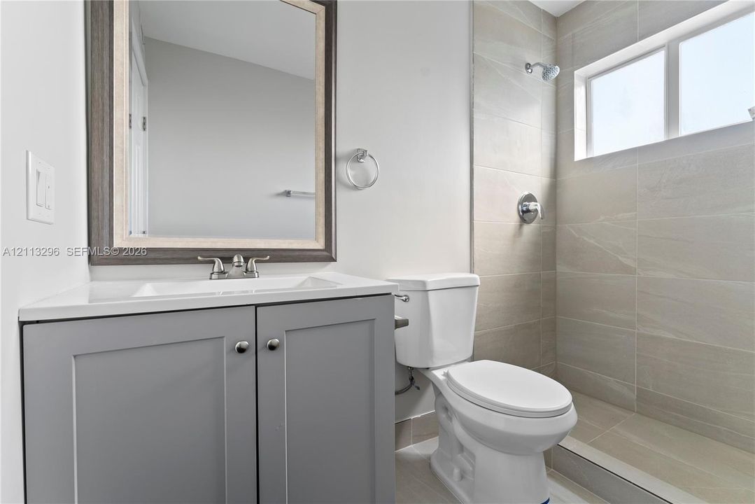 Bathroom featuring a gray shaker-style vanity with an integrated sink, framed wall-mounted mirror, ceramic tile shower surround, and a horizontal window