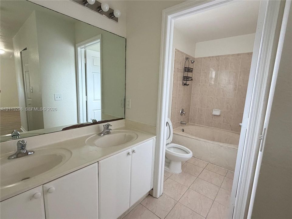 Dual-basin vanity with integrated sinks, large wall-mounted mirror, light-tone cabinetry, tiled flooring, and a combined tub and shower enclosure with wall-mounted ceramic tile surround