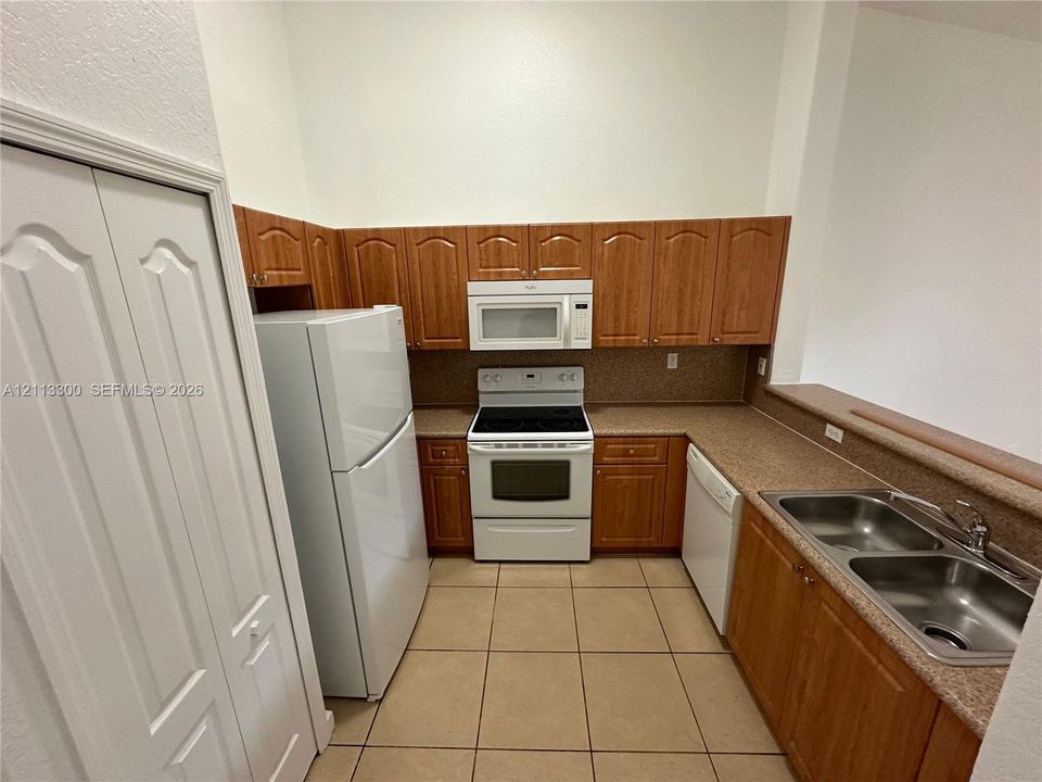 Kitchen featuring wood cabinetry, stone-finish countertops, tile flooring, dual-basin stainless steel sink, and white appliances